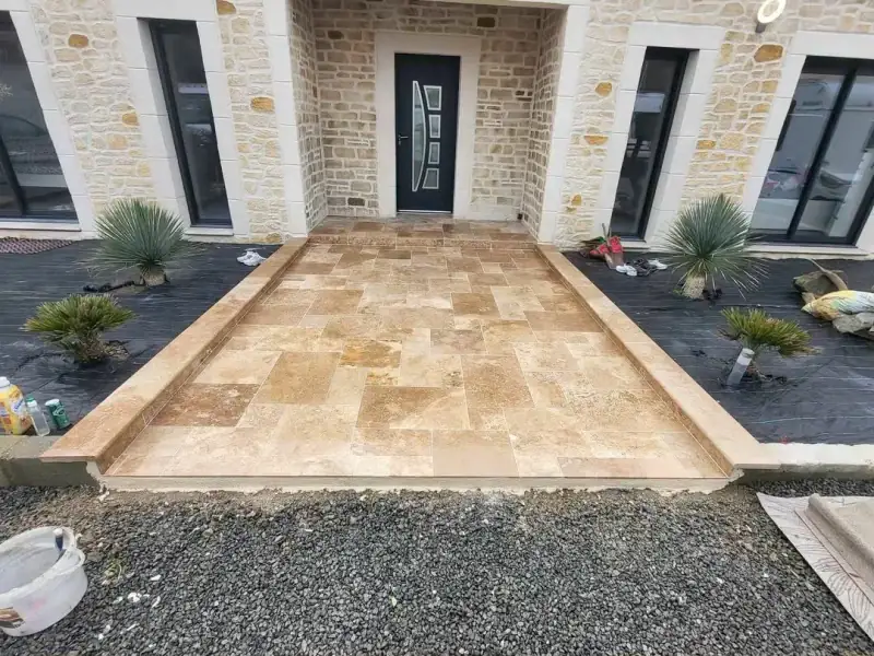 Terrasse d'entrée en dallage travertin avec margelles devant une maison à façade en pierre
